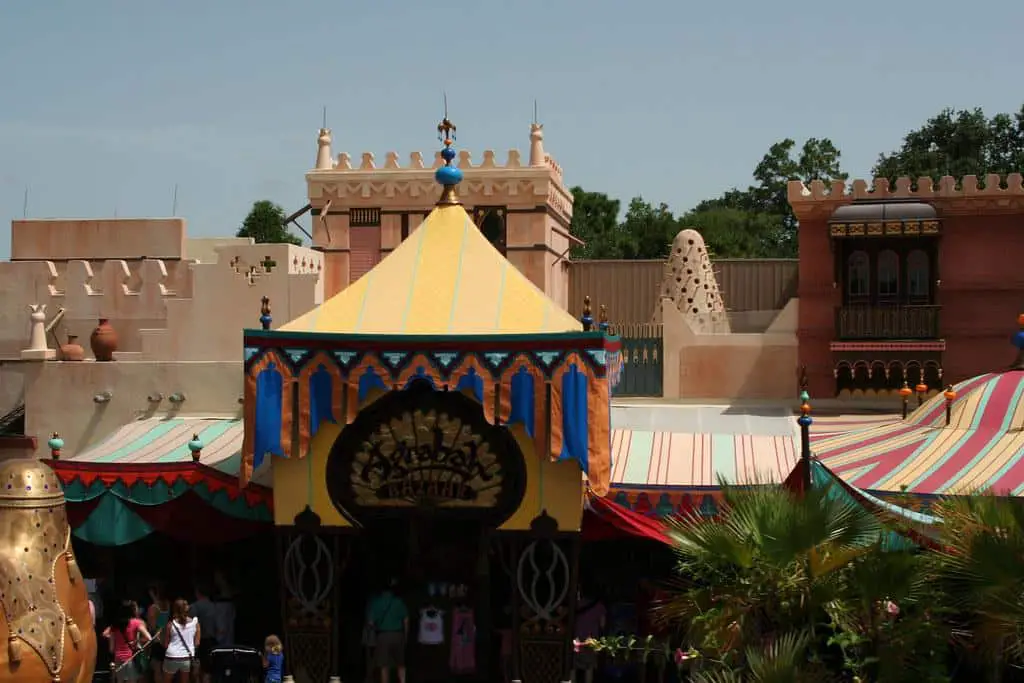 The bustling marketplace of Agrabah, with ornate buildings, colorful stalls, and a diverse crowd of Middle Eastern people